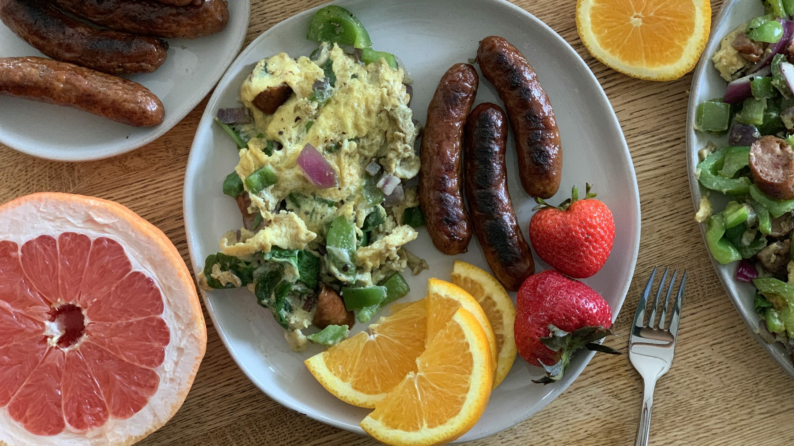 light grey plate with eggs and green pepper, charred sausage links, strawberries, orange slices, and half a grapefruit. One shiny fork. 