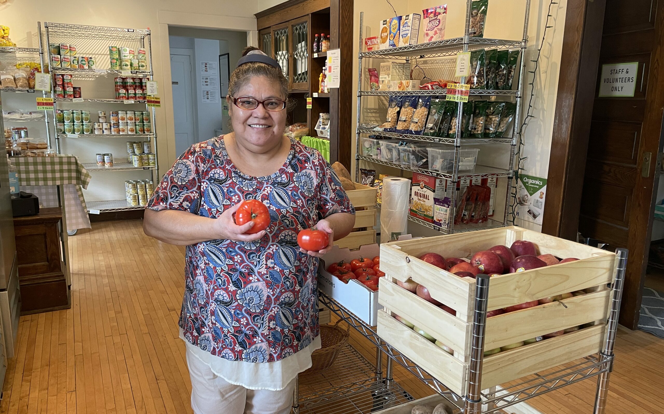 smiling person holding a bright heirloom tomato in each hand next to a wooden crate brimming with more!