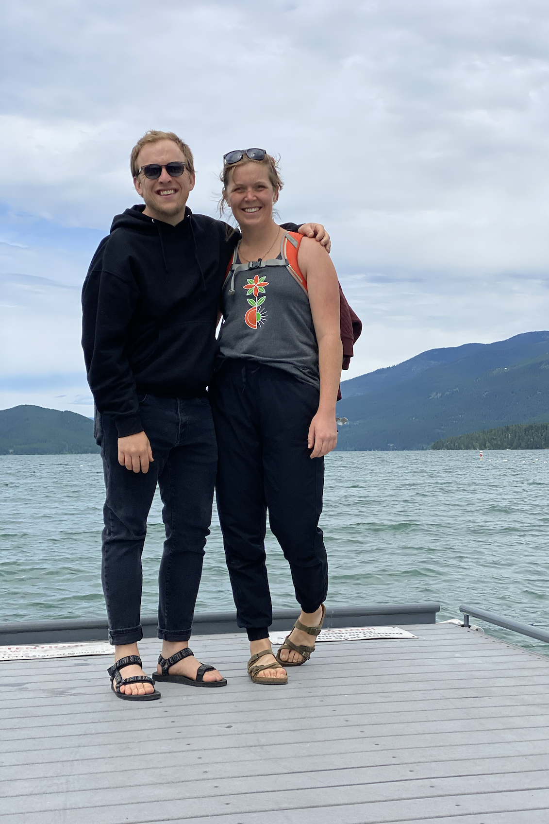 two people in black standing on a dock in front of some gorgeous aqua water with a little hill on the horizon